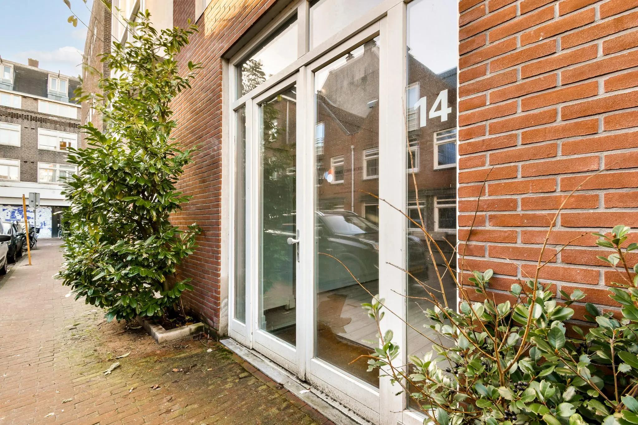 Glazen voordeur met huisnummer 14 aan de Korte Schimmelstraat, omringd door rode bakstenen en groene struiken.